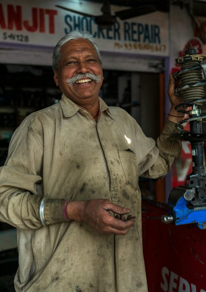 Cheerful elderly ethnic male worker in dirty uniform standing near equipment and looking at camera in workroom